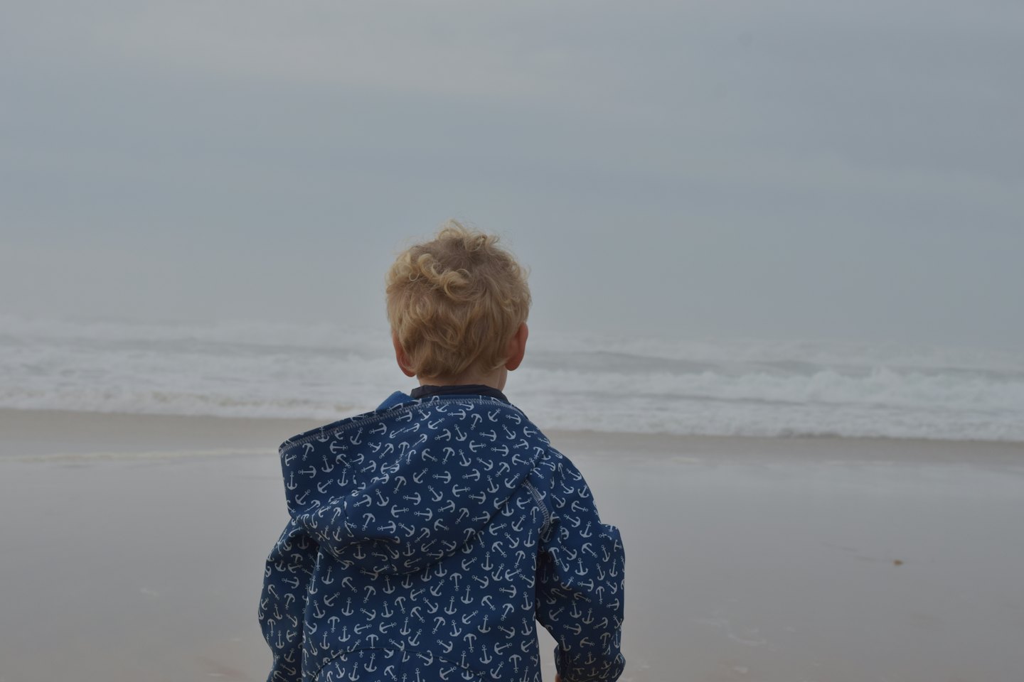 Kind in blauer Jacke steht am Strand und schaut auf die Wellen.